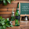 Reusable glass water bottle and stainless steel straw on a wooden table with 'Plastic-Free July' written on a chalkboard.